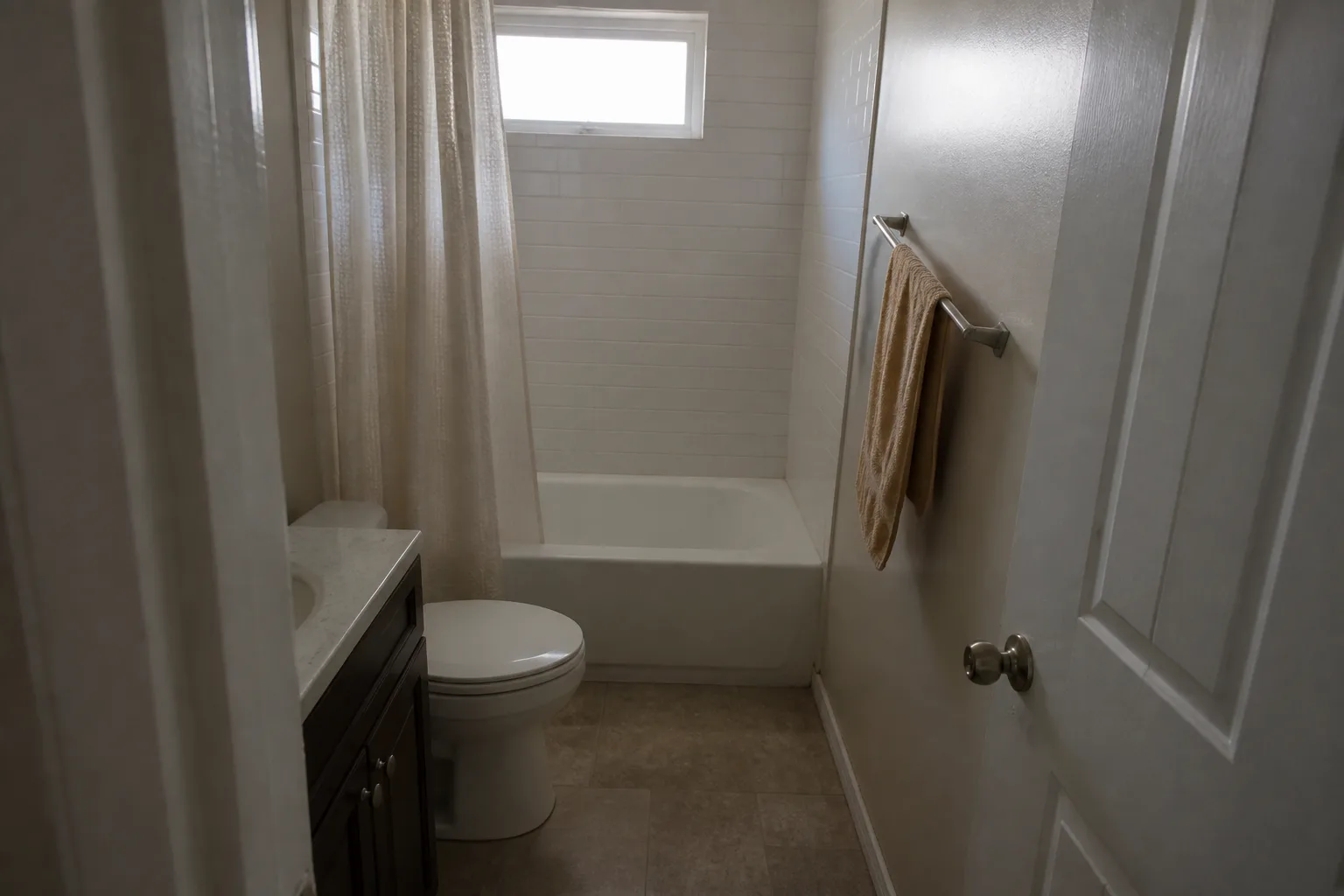 A small finished bathroom about five feet by eight, seen from the doorway: a tub with a tiled surround along the far wall, a toilet and one compact vanity tight together, lit by a single high window above the tub