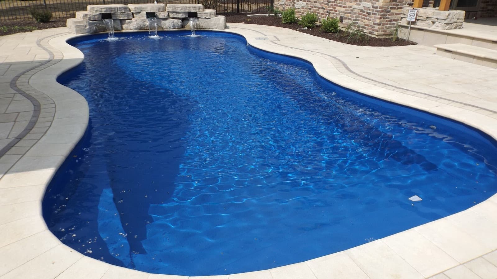 A blue free-form fiberglass pool with a stone water feature and light pavers